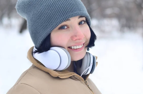 Oldukça çekici beyaz gözlü kız hipster kış park portresi. Kulaklıklar üzerinde müzik dinliyor. Gençlik sokak moda. Kış eğlence.