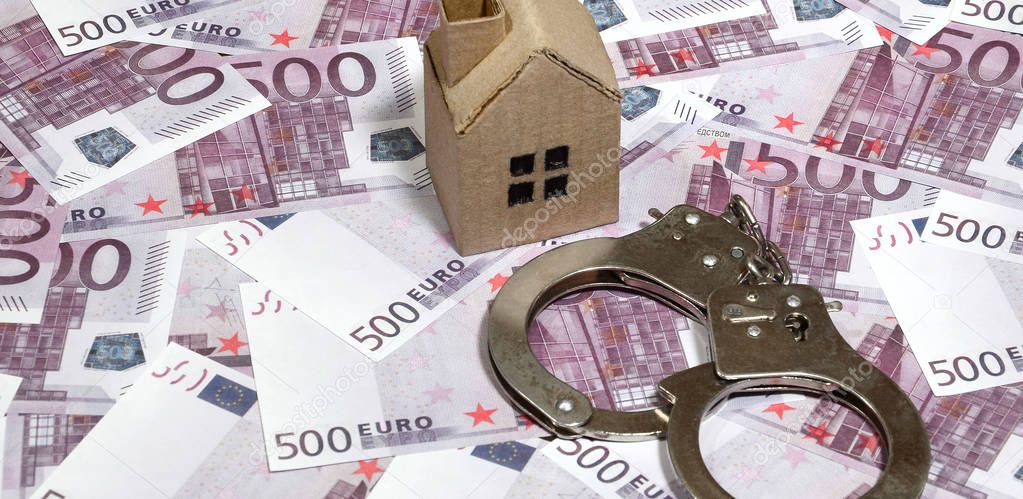 Scam with real estate. Cheating when buying or renting a house. Punishment for deception. A toy house with handcuffs on a many 500 purple euro banknotes