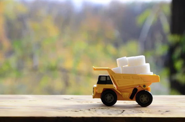 A small yellow toy truck is loaded with white sugar cubes. A car on a wooden surface against a background of autumn forest. Extraction and transportation of sugar