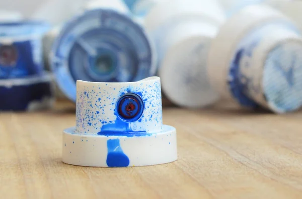 Several plastic nozzles from a paint sprayer that lie on a wooden surface against a gray wall background. The caps are smeared in blue paint. The concept of street art and graffiti