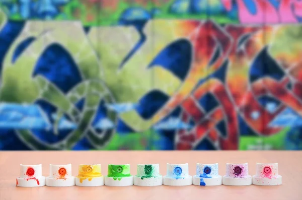 The soiled multicolored nozzles from the paint sprayer are lined up on a wooden table on a background of colored graffiti drawing in a wild style. The concept of street art