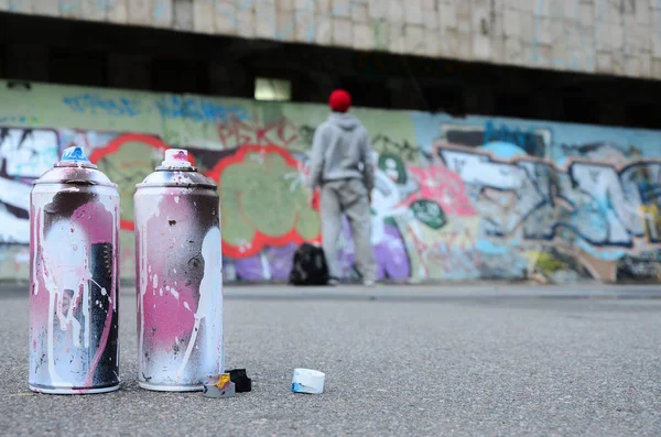 Several used spray cans with pink and white paint lie on the asphalt against the standing guy in front of a painted wall in colored graffiti drawings