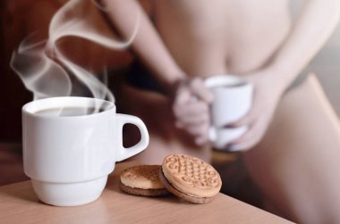 Bir fincan sıcak kahve ve yuvarlak kurabiye bir yatak üzerinde arka planda siyah iç çamaşırı seksi bir kız bir silueti ile. Ön plan öncelikli odak. Küçük beyaz kahve Kupası Buhar ve kahverengi bisküvi ile 