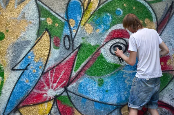 Photo of a young guy in denim shorts and a white shirt. The guy draws on the graffiti wall a drawing with aerosol paints of various colors. The concept of hooliganism and damage to property