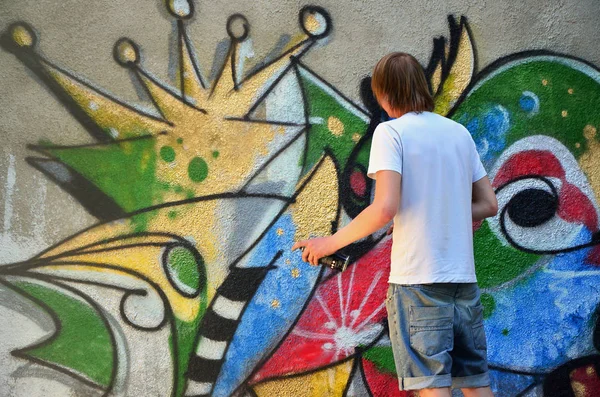 Photo of a young guy in denim shorts and a white shirt. The guy draws on the graffiti wall a drawing with aerosol paints of various colors. The concept of hooliganism and damage to property