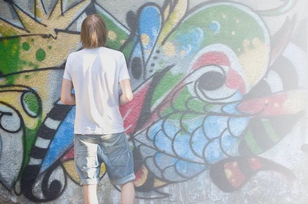 Photo of a young guy in denim shorts and a white shirt. The guy draws on the graffiti wall a drawing with aerosol paints of various colors. The concept of hooliganism and damage to property