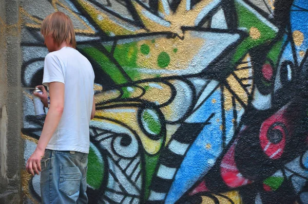 Photo in the process of drawing a graffiti pattern on an old concrete wall. Young long-haired blond guy draws an abstract drawing of different colors. Street art and vandalism concept