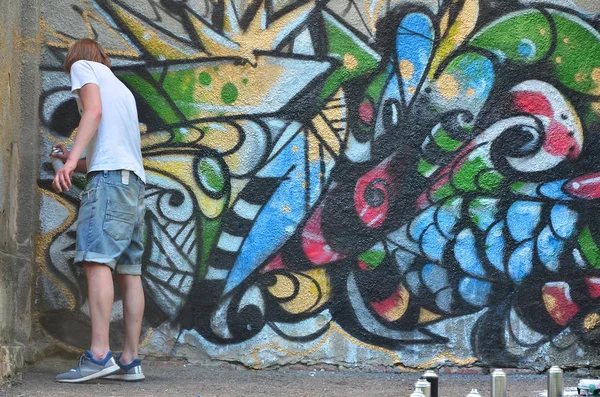 一个穿着牛仔短裤和白衬衫的年轻人的照片 那家伙在涂鸦墙上画了一幅带有各种颜色的气溶胶颜料的图画 流氓概念与财产损害 — 图库照片