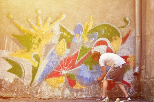 Photo of a young guy in denim shorts and a white shirt. The guy draws on the graffiti wall a drawing with aerosol paints of various colors. The concept of hooliganism and damage to property