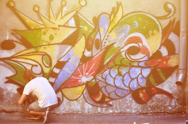 Photo of a young guy in denim shorts and a white shirt. The guy draws on the graffiti wall a drawing with aerosol paints of various colors. The concept of hooliganism and damage to property