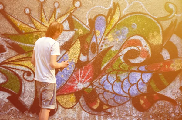 Photo of a young guy in denim shorts and a white shirt. The guy draws on the graffiti wall a drawing with aerosol paints of various colors. The concept of hooliganism and damage to property