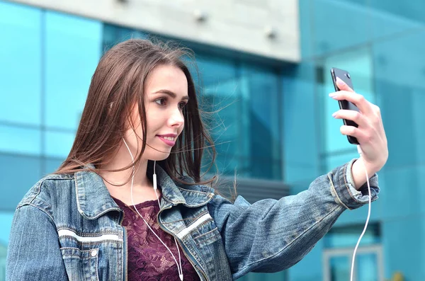年轻的黑发女孩穿着牛仔裤做智能手机办公大楼为背景拍照 时尚拍摄自己对手机相机的概念 — 图库照片