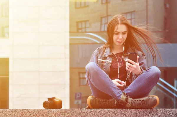 A young female photographer in jeans clothes uses a smartphone, sit on a granite parapet against a glazed office building. Concept for lessons or online photography learning and photo-training
