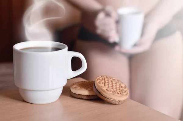 A cup of hot coffee and round cookies with a silhouette of a sexy girl in black underwear in the background on a bed. Focus foreground priority. Small white coffee cup with steam and brown biscuits