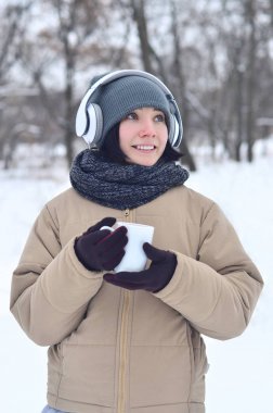 Güzel esmer beyaz genç kadın büyük beyaz kulaklık müzik ve içecek bir fincan kahve kış park veya ormandaki müzik dinlerken gülümseyen deldi