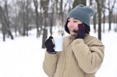 Oldukça çekici beyaz gözlü kız hipster kış Park portresi. Modern smartphone ve içecek kahve konuşuyor. Gençlik sokak moda. Açık havada yürüyüşler