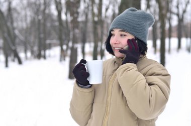 Oldukça çekici beyaz gözlü kız hipster kış Park portresi. Modern smartphone ve içecek kahve konuşuyor. Gençlik sokak moda. Açık havada yürüyüşler