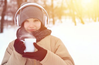 Güzel esmer beyaz genç kadın büyük beyaz kulaklık müzik ve içecek bir fincan kahve kış park veya ormandaki müzik dinlerken gülümseyen deldi