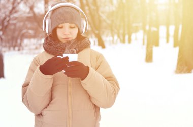 Güzel esmer beyaz genç kadın büyük beyaz kulaklık müzik ve içecek bir fincan kahve kış park veya ormandaki müzik dinlerken gülümseyen deldi