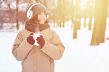 Güzel esmer beyaz genç kadın büyük beyaz kulaklık müzik ve içecek bir fincan kahve kış park veya ormandaki müzik dinlerken gülümseyen deldi
