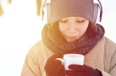 Güzel esmer beyaz genç kadın büyük beyaz kulaklık müzik ve içecek bir fincan kahve kış park veya ormandaki müzik dinlerken gülümseyen deldi