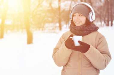 Güzel esmer beyaz genç kadın büyük beyaz kulaklık müzik ve içecek bir fincan kahve kış park veya ormandaki müzik dinlerken gülümseyen deldi