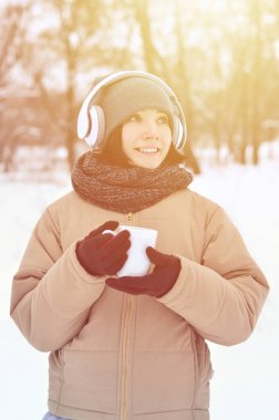 Güzel esmer beyaz genç kadın büyük beyaz kulaklık müzik ve içecek bir fincan kahve kış park veya ormandaki müzik dinlerken gülümseyen deldi