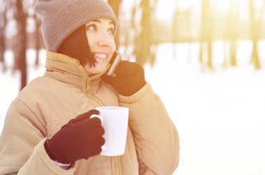 Oldukça çekici beyaz gözlü kız hipster kış Park portresi. Modern smartphone ve içecek kahve konuşuyor. Gençlik sokak moda. Açık havada yürüyüşler.