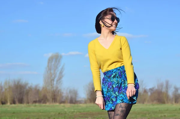 An attractive and pretty brunette in a bright yellow sweater and in round black glasses happily walks through the spring field. Photo of a joyful brunette during a spring sunny day