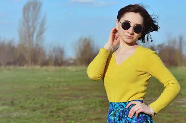 An attractive and pretty brunette in a bright yellow sweater and in round black glasses happily walks through the spring field. Photo of a joyful brunette during a spring sunny day