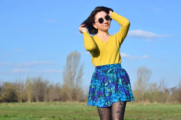 An attractive and pretty brunette in a bright yellow sweater and in round black glasses happily walks through the spring field. Photo of a joyful brunette during a spring sunny day