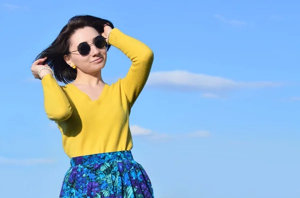 Portrait of an attractive and pretty brunette of European appearance in a bright yellow sweater and in round black glasses against a clear blue sky. Photo of a joyful brunette on a sunny day