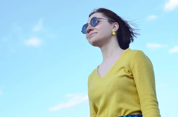 Portrait of an attractive and pretty brunette of European appearance in a bright yellow sweater and in round black glasses against a clear blue sky. Photo of a joyful brunette on a sunny day