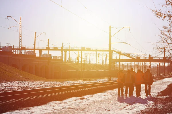 Several railway workers in signaling dirty orange uniforms are on the road next to the railway line. The train crew goes to work in the winter sunny morning.