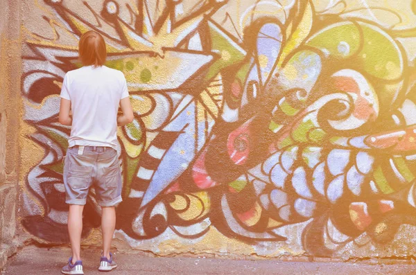 Photo of a young guy in denim shorts and a white shirt. The guy draws on the graffiti wall a drawing with aerosol paints of various colors. The concept of hooliganism and damage to property