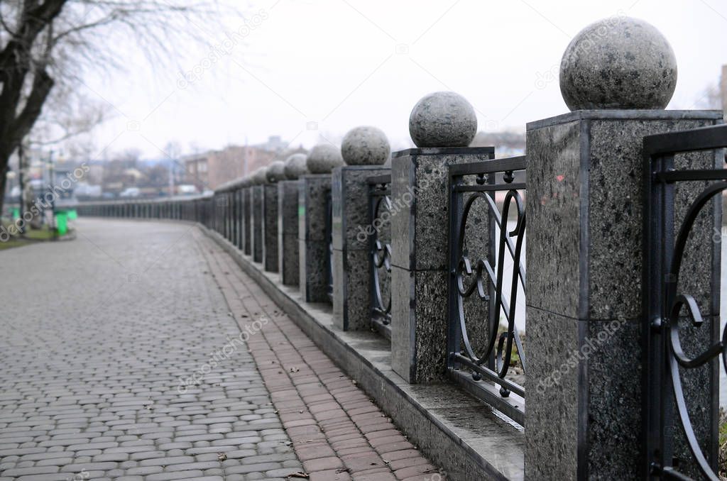 A beautiful granite fence with forged metal sections and decorative balls as decorations. The fence is built along the embankment of the street