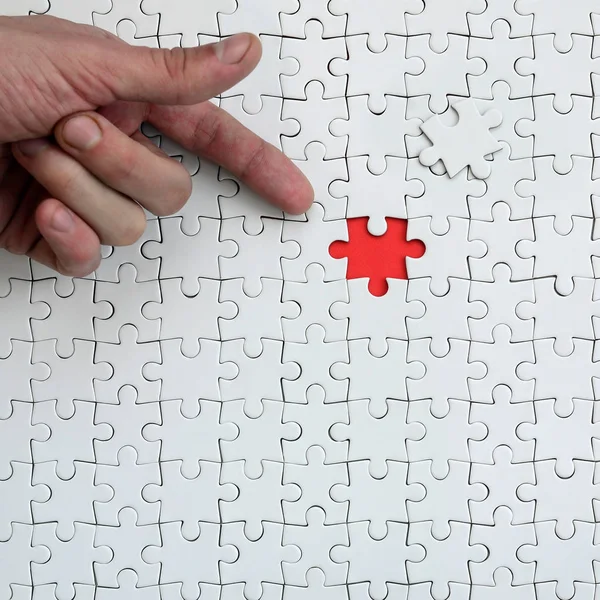 The texture of a white jigsaw puzzle in the assembled state with one missing element, forming a red space, pointed to by the finger of the male hand