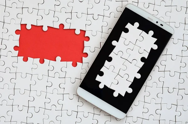 A modern big smartphone with several puzzle elements on the touch screen lies on a white jigsaw puzzle in an assembled state with missing elements