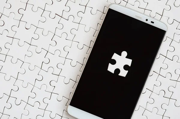 A modern big smartphone with several puzzle elements on the touch screen lies on a white jigsaw puzzle in an assembled state