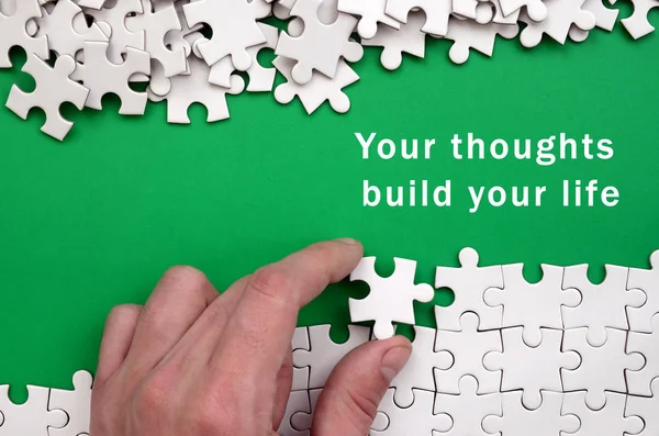 Your thoughts build your life. The hand folds a white jigsaw puzzle and a pile of uncombed puzzle pieces lies against the background of the green surface