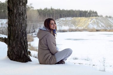 Kahverengi bir ceket genç bir beyaz kız kış döneminde bir ufuk çizgisi gökyüzü ve donmuş bir gölün arasında arka planda bir uçurum yakın oturuyor