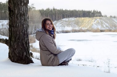 Kahverengi bir ceket genç bir beyaz kız kış döneminde bir ufuk çizgisi gökyüzü ve donmuş bir gölün arasında arka planda bir uçurum yakın oturuyor