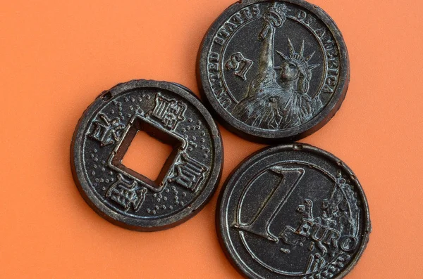 Three chocolate products in the form of Euro, USA and Japan coins lie on an orange plastic background. A model of cash coins in an edible form