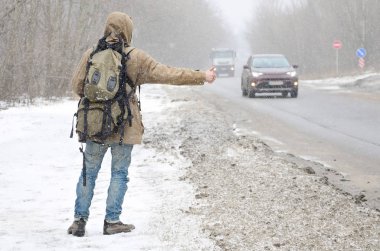 Bir büyük sırt çantası gösteren bir adamla bir banliyö asfalt yolda bir kış kar fırtınası içinde bir yolculuk sırasında otostop için başparmak