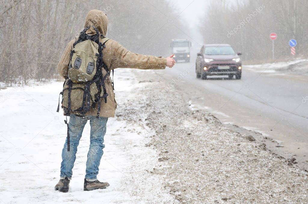 A man with a large backpack showing thumbs up for hitchhiking during road trip along a suburban asphalt road in a wintertime blizzard