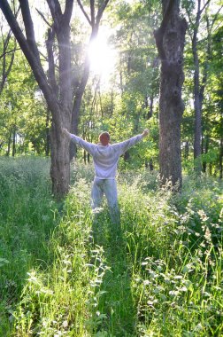 Gri spor elbiseli bir genç adam ormandaki ağaçlar arasında güneşin düşüyor sevinir. Rekreasyon spor sırasında açık hava ormanda çalıştırın. Dawn güzel zevk