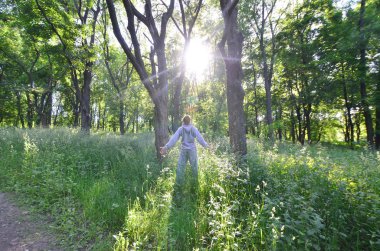 Gri spor elbiseli bir genç adam ormandaki ağaçlar arasında güneşin düşüyor sevinir. Rekreasyon spor sırasında açık hava ormanda çalıştırın. Dawn güzel zevk