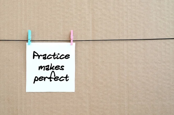 Practice makes perfect. Note is written on a white sticker that hangs with a clothespin on a rope on a background of brown cardboard