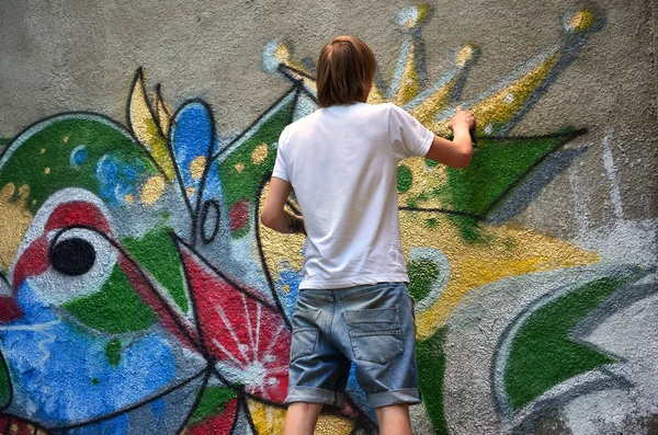 Photo of a young guy in denim shorts and a white shirt. The guy draws on the graffiti wall a drawing with aerosol paints of various colors. The concept of hooliganism and damage to property