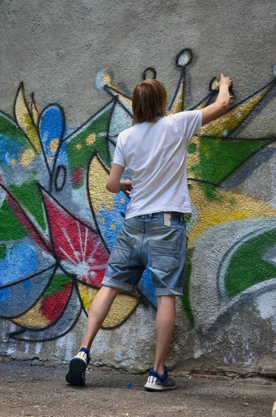 Photo of a young guy in denim shorts and a white shirt. The guy draws on the graffiti wall a drawing with aerosol paints of various colors. The concept of hooliganism and damage to property